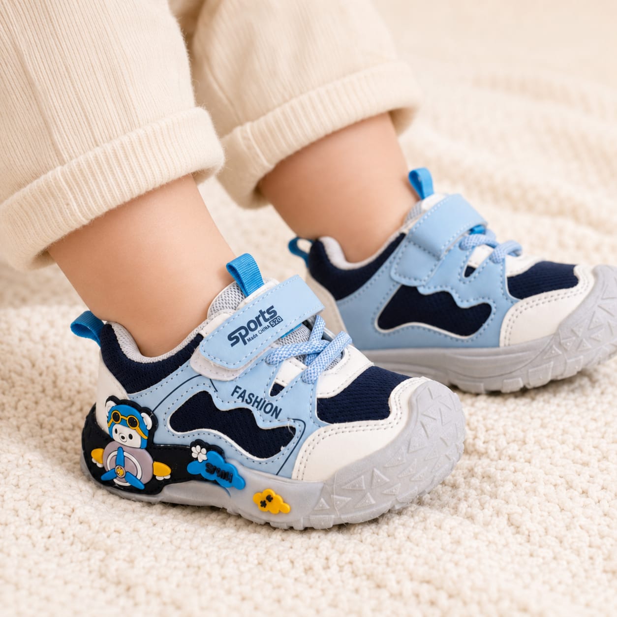 A pair of toddler's light blue and navy sneakers with white accents, shown on a child's feet wearing beige pants on a soft cream-colored carpet. The shoes feature hook-and-loop straps, a decorative cartoon bear on the side, and a textured sole.