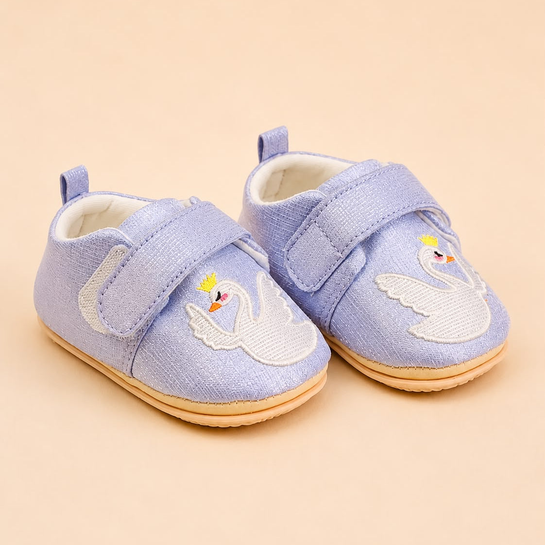 A pair of baby shoes in light blue canvas with embroidered swans on the toes, featuring hook-and-loop straps and a natural rubber sole, set against a solid beige background.
