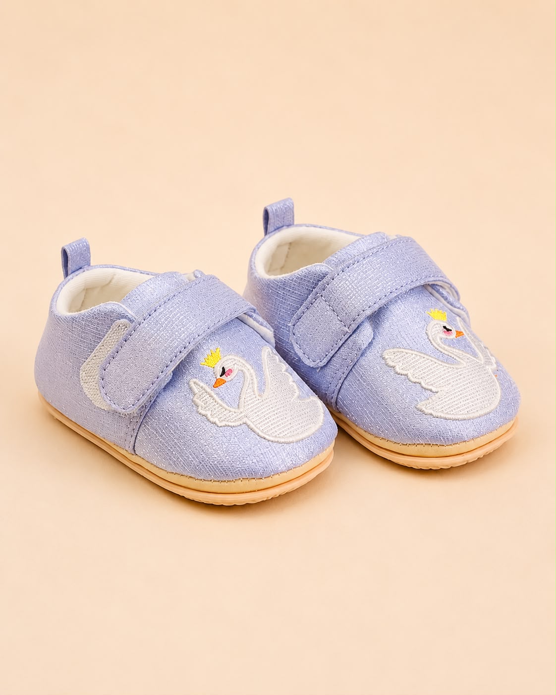 A pair of baby shoes in light blue canvas with embroidered swans on the toes, featuring hook-and-loop straps and a natural rubber sole, set against a solid beige background.